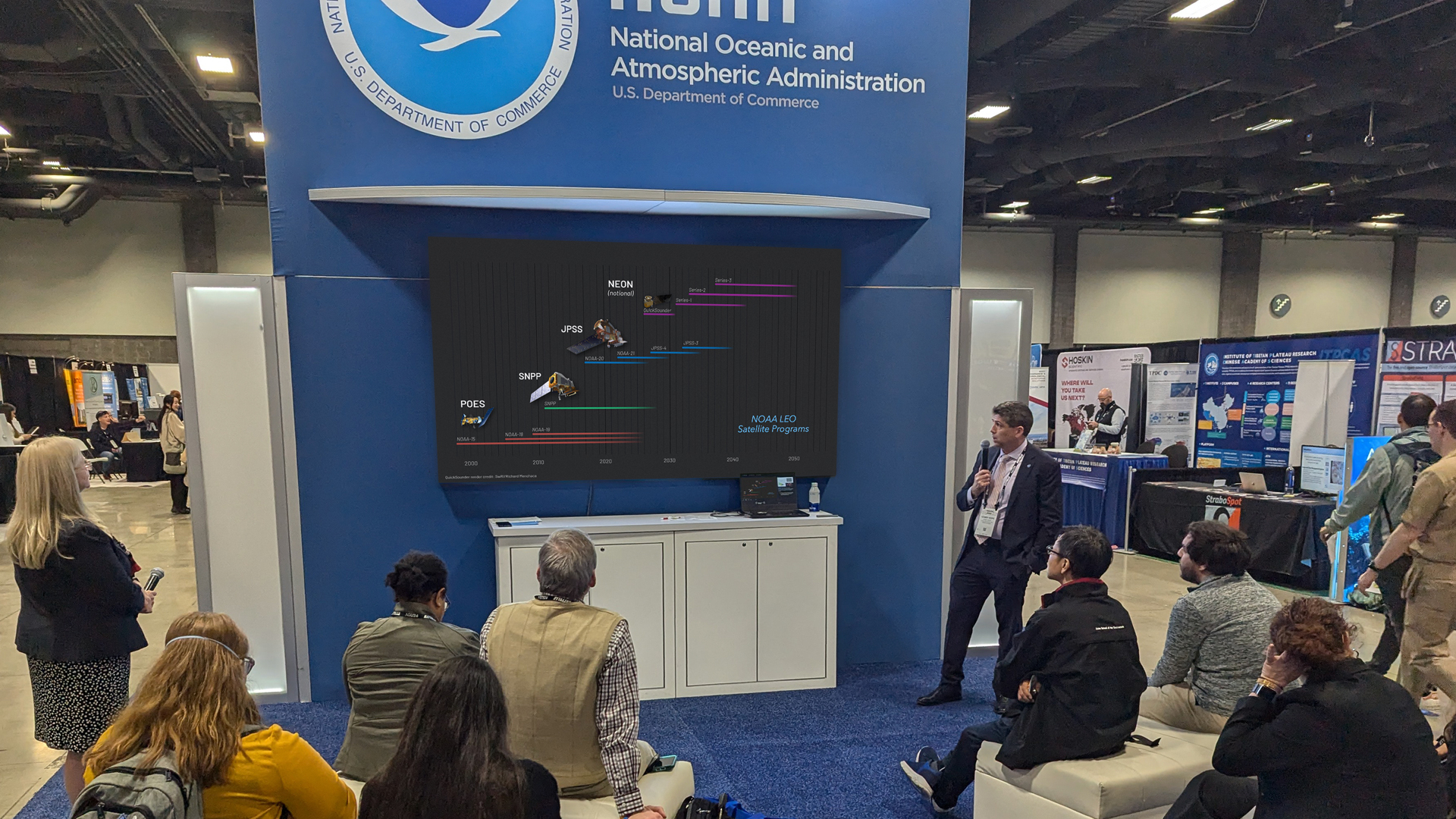 At a conference booth structure with the NOAA signage, a presenter looks at a large screen while a group of people sit in front watching the presentation.