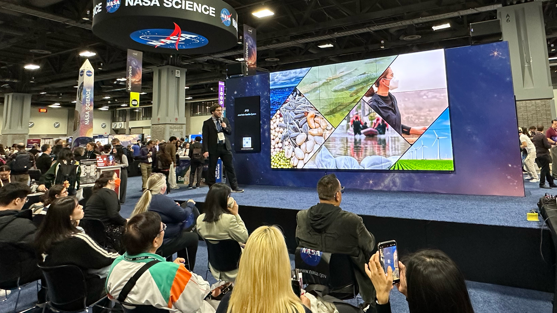 At a conference exhibit hall, a man on a stage stands next a large multi-panel screen. A crowd sits watching as he presents. People are seen in the background mingling on the exhibit hall.