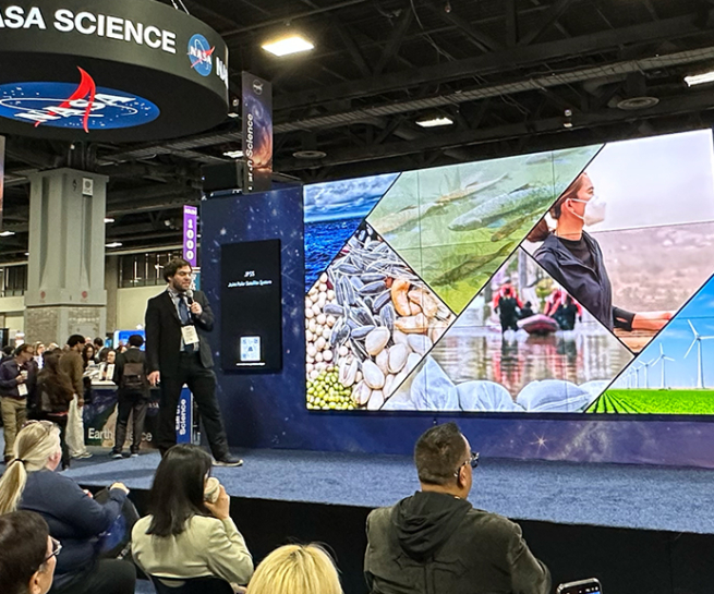 At a conference exhibit hall, a man on a stage stands next a large multi-panel screen. A crowd sits watching as he presents. People are seen in the background mingling on the exhibit hall.